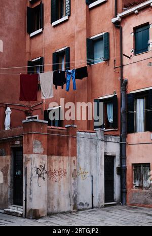 Gedämpftes rotes Wohngebäude mit grünen Fensterläden. Waschen hängt an der Schlange, die vom Fenster gestreckt ist, Venedig, Italien, mehrere Eingänge Stockfoto