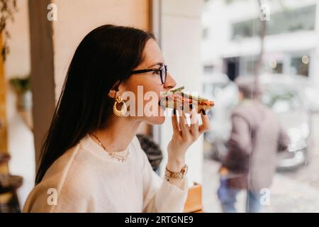 Seitenansicht der hübschen jungen Frau im stilvollen Outfit beißen Leckeres Sandwich, während man in einem gemütlichen Café neben dem Fenster sitzt Stockfoto