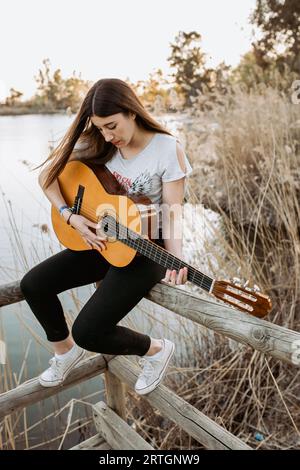 Ziemlich junge Frau in casual Outfit Gitarre spielen, während sitzen Am Holzzaun in der Nähe von ruhigen See an sonnigen Tag Stockfoto