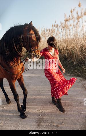 dame in gepunkteten roten Kleid führt tänzelnden Pferd auf dem Weg In der Nähe der Wiese Stockfoto