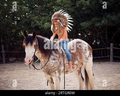 Junger Jockey mit Federkopfbedeckung, der zu Pferd mit Zügen sitzt, während er in der Nähe von Zäunen und grünen Bäumen auf das Pferd schaut Stockfoto