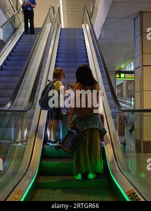 Rückansicht von Frau und Jungen mit Gepäck, das auf die Abfahrt am Flughafen-Terminal wartet und einem Mann, der die Rolltreppe herunterfährt Stockfoto