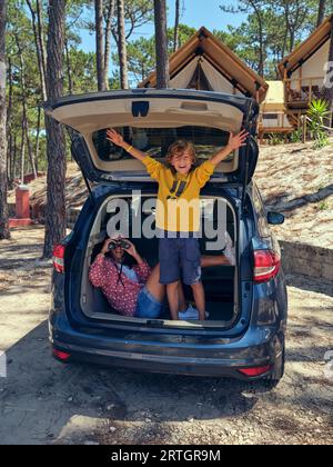 Eine ganze Länge von Frau, die durch ein Fernglas schaut, und lächelnder Junge, der im Kofferraum des Autos steht, der in der Nähe von Glamping-Hütten geparkt ist, die während der Sommerreise in den Wald gelegt werden Stockfoto