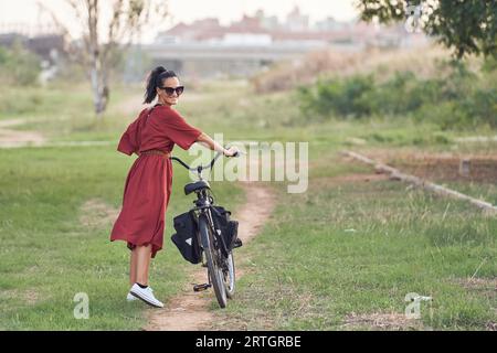 Seitenansicht einer jungen Radfahrerin in rotem Maxikleid und Sonnenbrille, die mit dem Fahrrad auf dem Weg spaziert, während sie lächelt und über die Schulter schaut Stockfoto