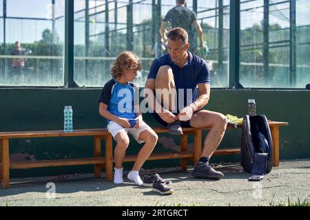 Ein ganzer Körper von ernsthaften Männern und Jungen, die auf einer Bank auf der Straße sitzen, während sie sich auf das Training mit Padelball und Schlägern vorbereiten Stockfoto