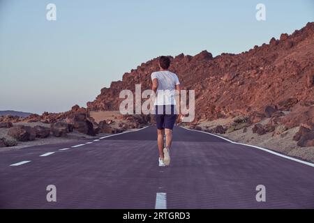 Ganzkörper-Rückansicht eines anonymen aktiven Mannes, der am Sommertag auf einer leeren Asphaltstraße in der Nähe von Bergrücken in der Natur läuft Stockfoto