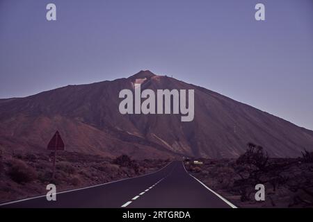 Gerade, leere Asphaltstraße mit Markierungslinien unter wolkenlosem Himmel, die morgens in Spanien zum rauen Teide-Vulkan in der Natur führt Stockfoto