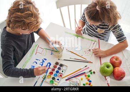Hoher Winkel von Bruder und Schwester, die Pinsel halten und Bilder auf weißen Papierbögen mit Aquarellfarbe zeichnen Stockfoto