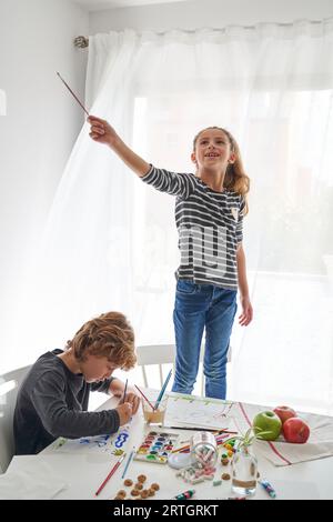 Lächelndes Mädchen, das auf einem Stuhl steht und Pinsel hält, und Junge, der auf einem Papierblatt mit Aquarellfarben zeichnet, sitzt am Tisch mit verschiedenen Süßigkeiten darauf Stockfoto