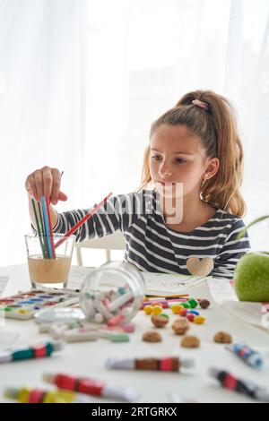 Konzentriertes Mädchen, das Pinsel in Glas mit Wasser wählt, während es am unordentlichen Tisch mit verstreuten Buntstiften und Farben in hellem Raum malt Stockfoto