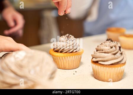 Ernten Sie anonyme Köchin, die cremige Muffins mit geriebener Schokolade an der Theke in der Küche bestreut Stockfoto