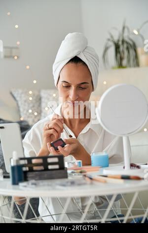 Erwachsene, fokussierte Frau mit Handtuch auf dem Kopf, die Lidschatten auf der Hand aufträgt, während sie morgens Make-up macht und Schönheitsblogs auf dem Mobiltelefon aufnimmt Stockfoto