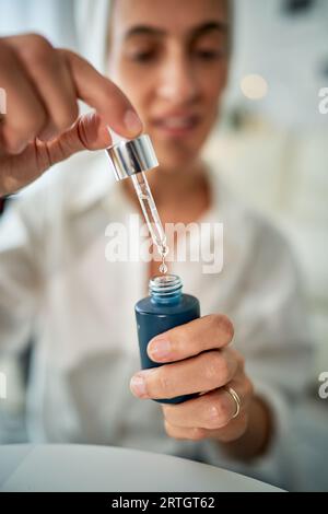 Crop Uncrered female Show bottle with Fluid Facial Moisturizing Serum with Pipette and Drop of Water while doing Skincare Routine in morning Stockfoto