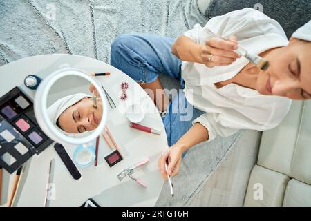 Über dem Kopf einer Frau, die auf dem Boden sitzt und Hautpflegeverfahren durchführt, die kosmetische Feuchtigkeitsmaske mit dem Pinsel auf das Gesicht aufträgt Stockfoto