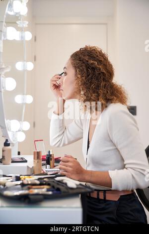 Seitenansicht einer jungen lockigen Frau, die Augen mit Kosmetikprodukten schminkt, die vor einem beleuchteten Spiegel sitzen Stockfoto