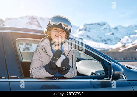 Begeistert war der Junge in der Skibrille, der die Kamera betrachtete, während er sich während des Roadtrips am Wintertag aus dem Autofenster mit Schneeball lehnte Stockfoto