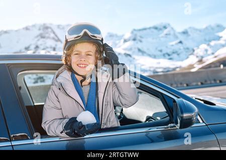 Positiver Junge in Skibrille, der wegschaut, während er sich mit Schneebalg beim Roadtrip gegen verschneite Berge aus dem Autofenster lehnt Stockfoto