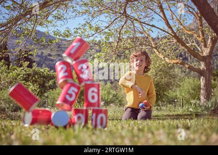 Positiver Junge kniend auf grasbewachsener Wiese und wirft Säcke in Pyramiden aus roten Blechdosen, während er in der Natur spielt Stockfoto