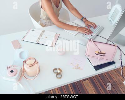 Von oben nach unten eine unerkennbare Frau, die mit Notizbüchern am weißen Tisch sitzt und während der Arbeit im Büro auf der Tastatur des Computers schreibt Stockfoto