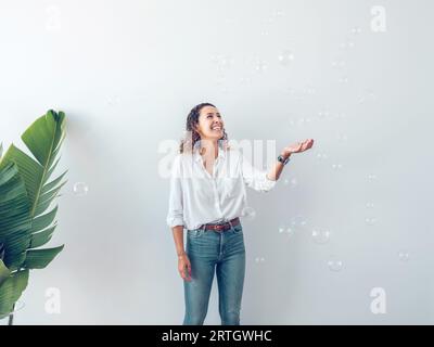 Fröhliche junge Frau in lässiger Kleidung, die sich freut und überraschend Seifenblasen fallen lässt, während sie an einer weißen Wand in der Nähe einer grünen Pflanze steht Stockfoto