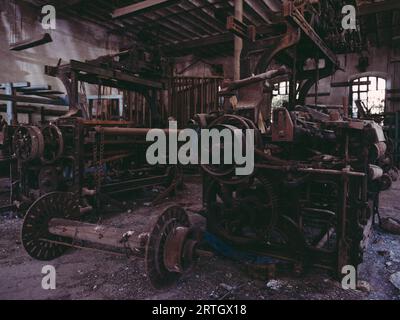 Alte, rostige Metallwebstühle mit Zahnrädern und defekten Geräten, die auf beschädigtem Boden in einer ausrangierten Industriegarnfabrik platziert wurden Stockfoto