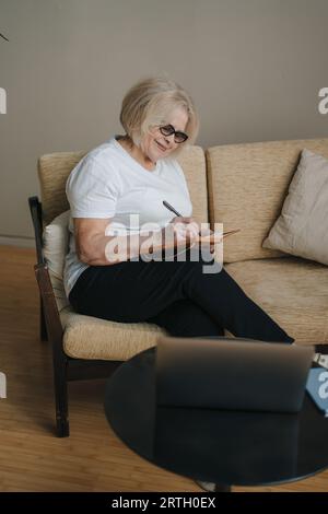 Porträt der lächelnden älteren Frau mit Laptop, der online arbeitet und Notizen macht, die zu Hause auf dem Sofa sitzen. Freiberufliche Karriere und Lifestyle. Lässig Stockfoto