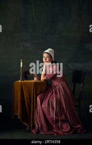 Hübsche, träumende junge Frau im Bild eines Renaissance-Dienstmädchen, das am Tisch sitzt und einen Brief vor dunklem Vintage-Hintergrund schreibt Stockfoto