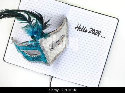 Notizbuch mit „Hello 2024“-Schriftzug und Karnevalsmaske auf weiß Stockfoto