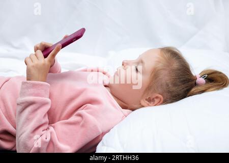 Kleines Mädchen (3-4 Jahre) liegt im Bett und schaut auf den Smartphone-Bildschirm. Das Kind schaut sich Karikaturen an, spielt Spiele vor einem Traum. Stockfoto