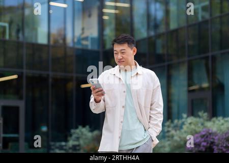 Erfolgreicher asiatischer Mann in zwangloser Kleidung benutzt das Telefon, Mann geht in der Stadt in der Nähe eines modernen Gebäudes spazieren, wählt eine Nachricht, ruft an, Geschäftsmann nach der Arbeit. Stockfoto
