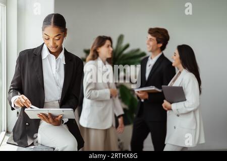 afroamerikanische Chefin, die ein digitales Tablet verwendet und im Büro mit einer Gruppe verschiedener Kollegen im Hintergrund arbeitet Stockfoto