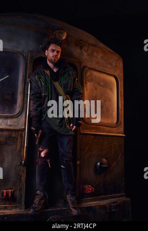 Postapokalyptischer Überlebender in getragener Kleidung, der mit einer Waffe auf einem rostigen U-Bahn-Wagen im Untergrund steht Stockfoto