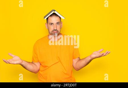 Bärtiger hispanischer Mann in seinen 40ern, der ein oranges T-Shirt trägt, ein offenes Buch auf dem Kopf hält und seine Hände hebt, da er nichts versteht Stockfoto