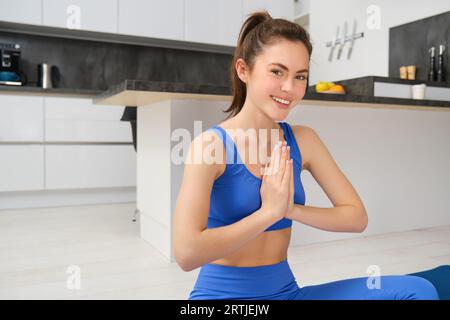 Junge Frau, Fitnesstrainer macht Yoga von zu Hause aus, schließt Hände zusammen, nameastische Geste, sitzt mit überkreuzten Beinen auf einer Gummimatte und meditiert von zu Hause aus Stockfoto