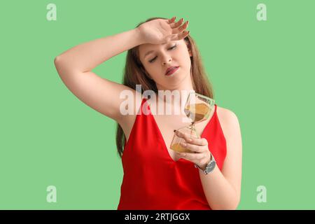 Müde junge Frau mit Sanduhr auf grünem Hintergrund. Terminkonzept Stockfoto