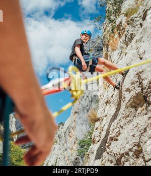 Teenager Junge auf der natürlichen Klippe Kletterwand. Der Junge hing mit einem Klettergurt an einem Seil und der Vater brachte ihn mit einem bela auf den Boden Stockfoto