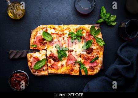 Heiße hausgemachte römische Pizza mit Schinken, Mozzarella-Käse, sonnengetrockneten Tomaten, Sauce und grünem Basilikum, schwarzer Tischhintergrund, Blick von oben Stockfoto