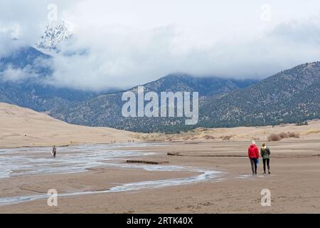 Ein Paar spaziert im Frühjahr im Great Sand Dunes National Park flussaufwärts in Richtung der weit entfernten schneebedeckten Sangre de Cristo Mountains Stockfoto