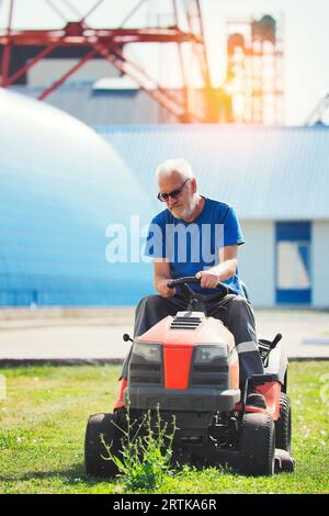 Arbeiter mäht das Gebiet der Industrieanlage. Man fährt an sonnigen Sommertagen selbstfahrenden Rasenmäher auf dem Rasen. Ältere Rentnermondlichter als Gärtner. Stockfoto