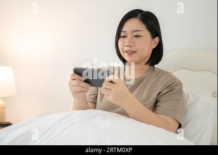 Eine schöne und glückliche asiatische Plus-Size-Frau in bequemer Kleidung spielt gerne mobile Spiele auf ihrem Smartphone, während sie sich auf ihrem Bett in ihrem Schlafzimmer entspannt. P Stockfoto