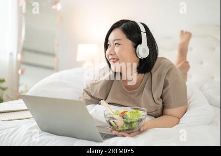 Eine glückliche asiatische Plus-Size-Frau liegt auf dem Bett und genießt ihren gesunden Salat, während sie sich einen Film auf ihrem Laptop in ihrem Schlafzimmer ansieht. Stockfoto