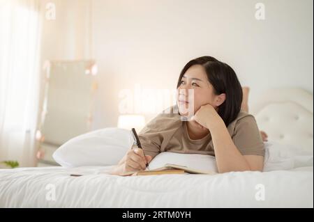 Eine charmante und glückliche asiatische Plus-Size-Frau träumt von ihrem Glück und Erfolg, während sie ihr Tagebuch führt oder etwas in ihr Notizbuch schreibt Stockfoto