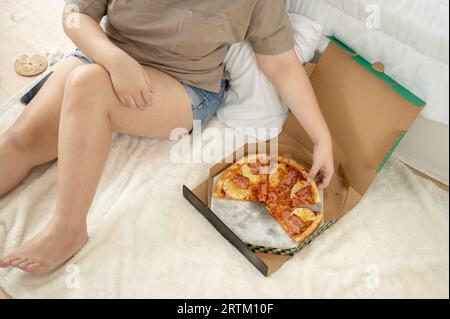 Abgeschnittenes Bild von oben: Eine kurvige Frau in Übergröße in bequemer Kleidung, die am Wochenende Pizza in ihrem Schlafzimmer isst. Ungesunder Lebensstil, häusliches Leben, jun Stockfoto