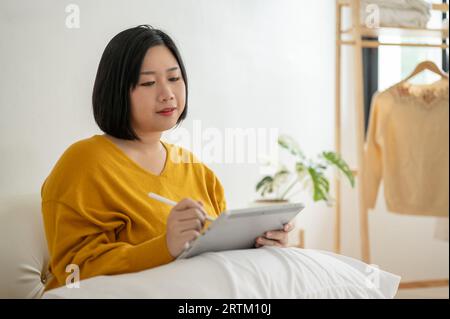 Eine schöne und glückliche asiatische Plus-Size-Frau in lässiger Kleidung benutzt ihr digitales Tablet auf einem Sofa in ihrem Wohnzimmer. Menschen- und Technologiekonzepte Stockfoto