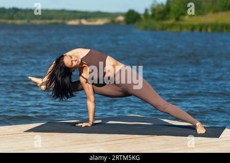 Frau, die Yoga praktiziert, Ardha Visvamitrasana-Übung durchführt, Salbei-Pose, allein in Sportbekleidung am Ufer eines Sees an einem sonnigen Tag trainiert Stockfoto