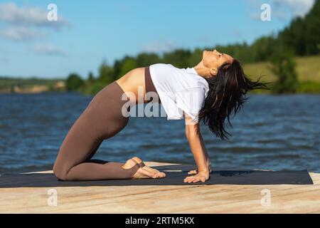 Eine Frau macht Yoga, führt Ardha Chandrasana-Übungen durch, hält eine halbe Brücke und trainiert an einem sonnigen Morgen alleine in Sportkleidung am Ufer eines Sees Stockfoto