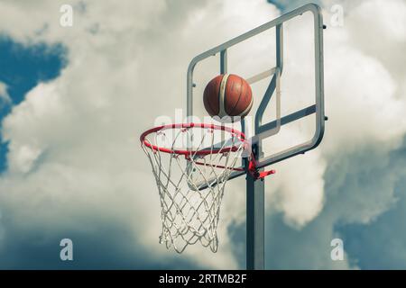 Der orangefarbene Basketballball tritt in den Basketballkorb ein. Spielen auf der Straße auf dem Outdoor-Court mit einem dramatischen Himmel und Wolken im Hintergrund, nah Stockfoto