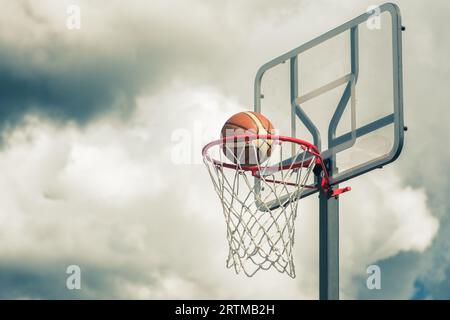 Der orangefarbene Basketballball tritt in den Basketballkorb ein. Spielen auf der Straße auf dem Outdoor-Court mit einem dramatischen Himmel und Wolken im Hintergrund, nah Stockfoto