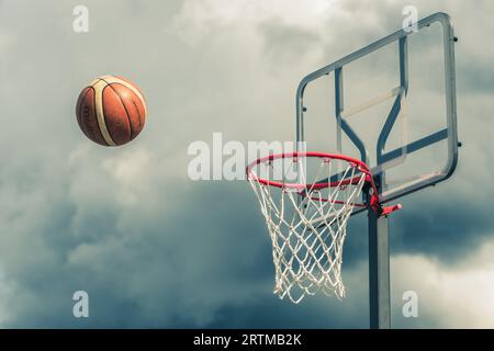 Der orangefarbene Basketballball tritt in den Basketballkorb ein. Spielen auf der Straße auf dem Outdoor-Court mit einem dramatischen Himmel und Wolken im Hintergrund, nah Stockfoto