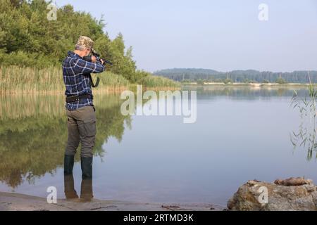 Mann mit Jagdgewehr in der Nähe des Sees im Freien. Leerzeichen für Text Stockfoto